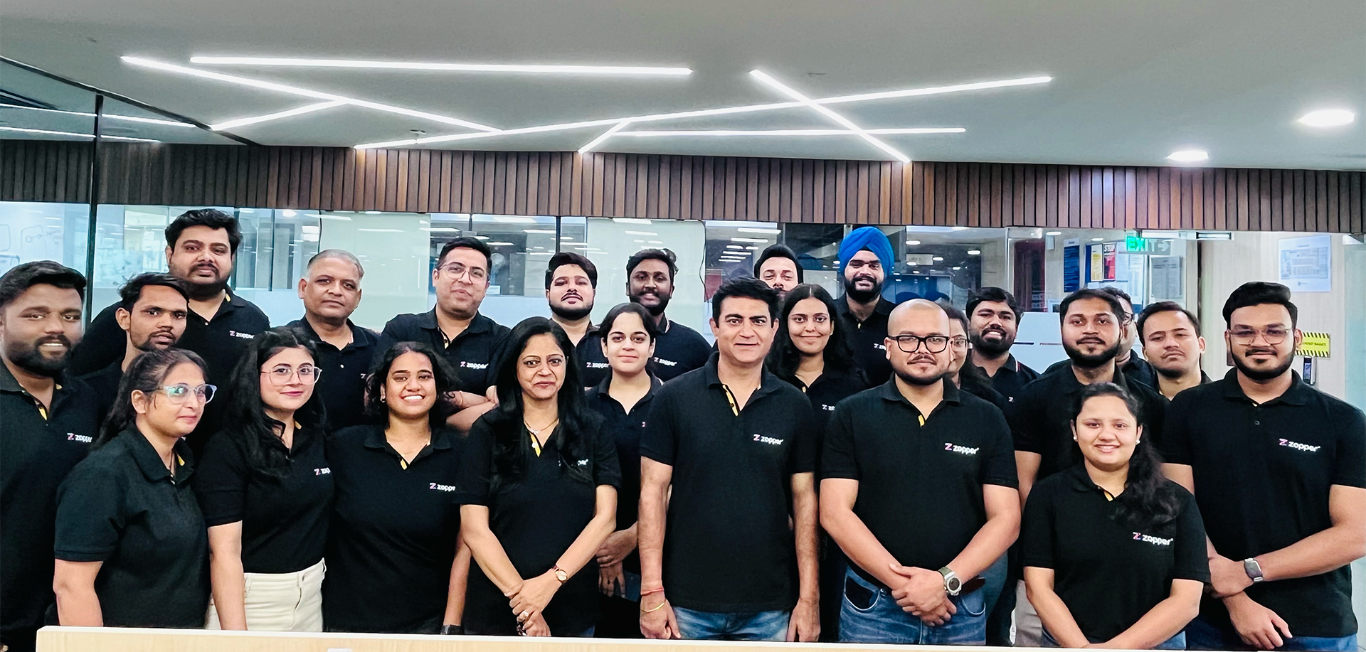 Group of smiling men and women wearing black Zopper polo shirts posing indoors under modern ceiling lights.