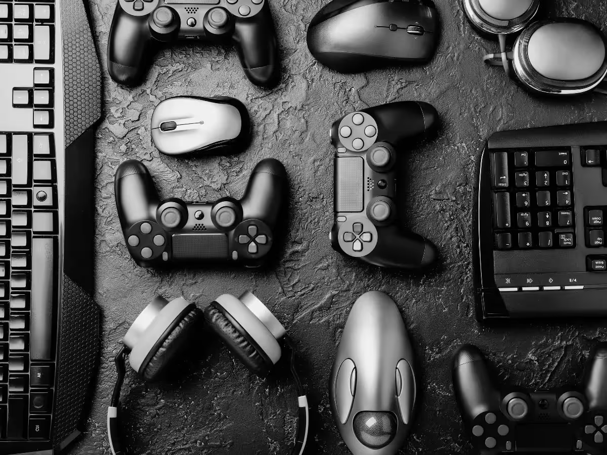 Czarno-białe zdjęcie typu flat lay przedstawiające akcesoria gamingowe: pady, myszki komputerowe, słuchawki i klawiatury na ciemnym, teksturowanym tle