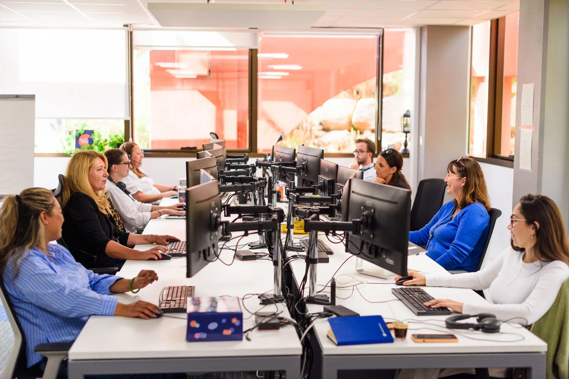 équipe du support client Preventimmo travaillant sur ordinateurs dans un bureau lumineux avec grandes fenêtres.
