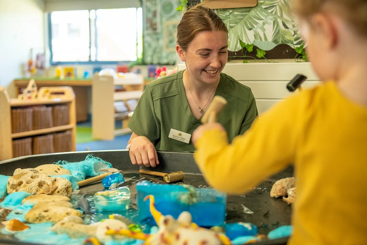 A nursery teacher smiles as a child plays with water, ice, and toys at a sensory table.