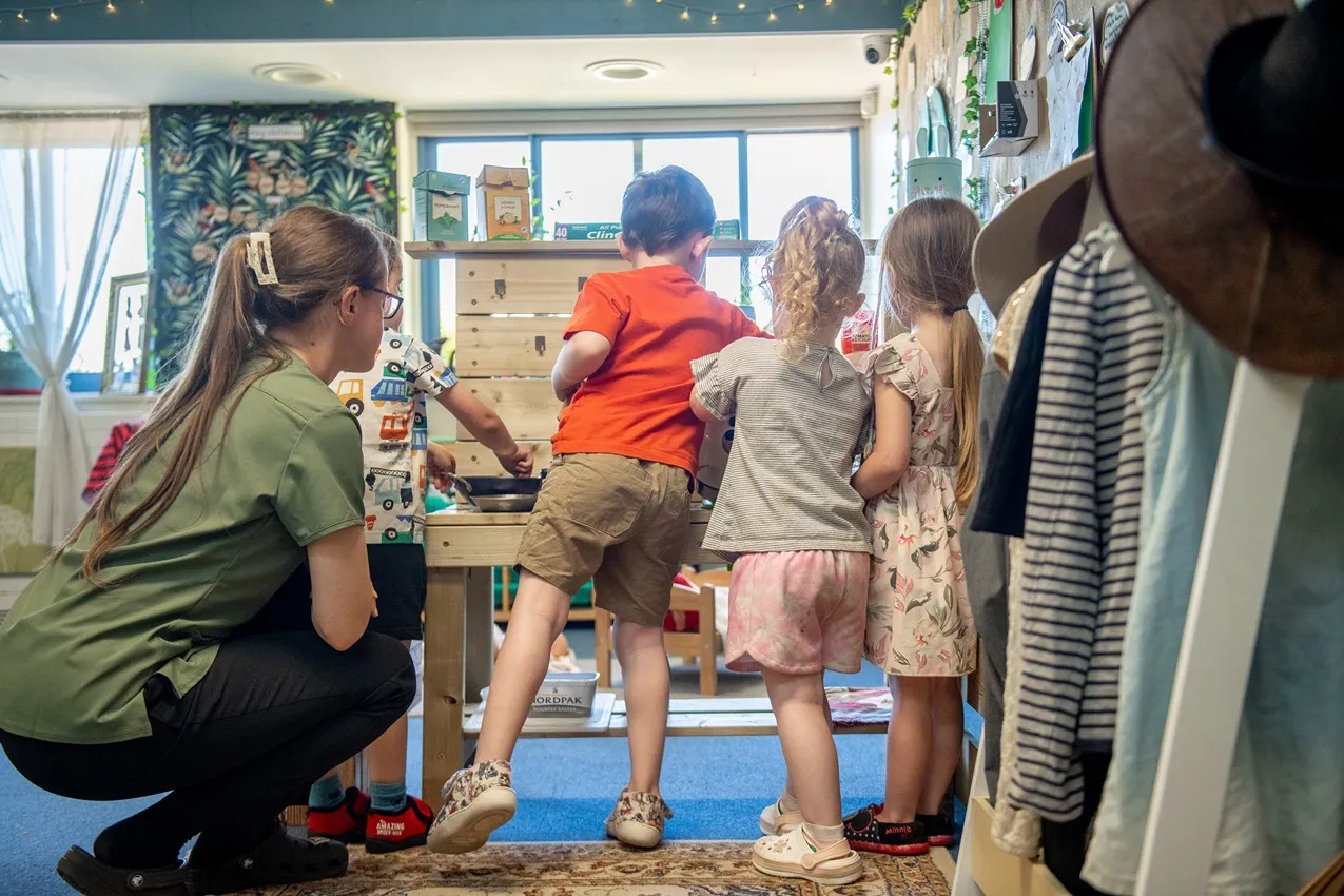 Children playing at a wooden play kitchen with a teacher in a bright nursery setting.