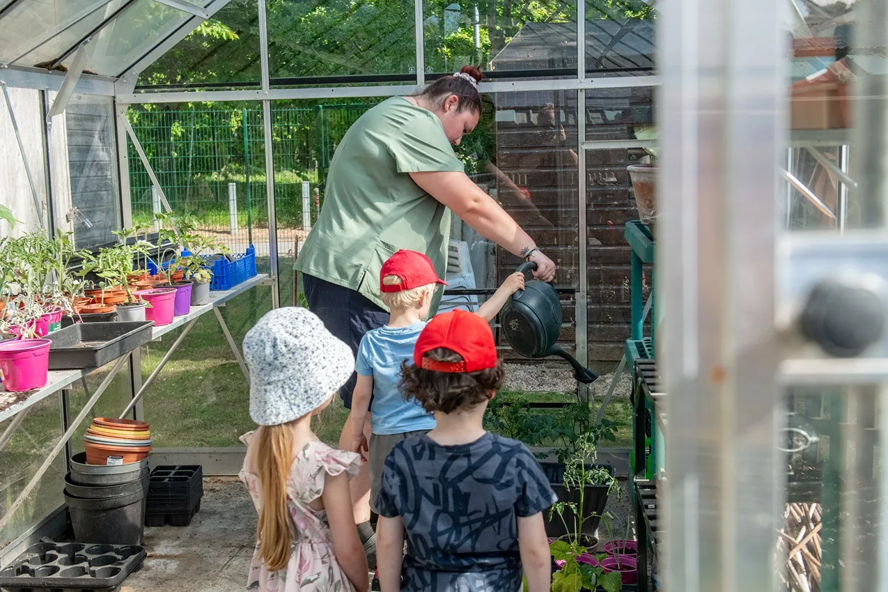 A woman watering plants in a greenhouse with three children watching.
