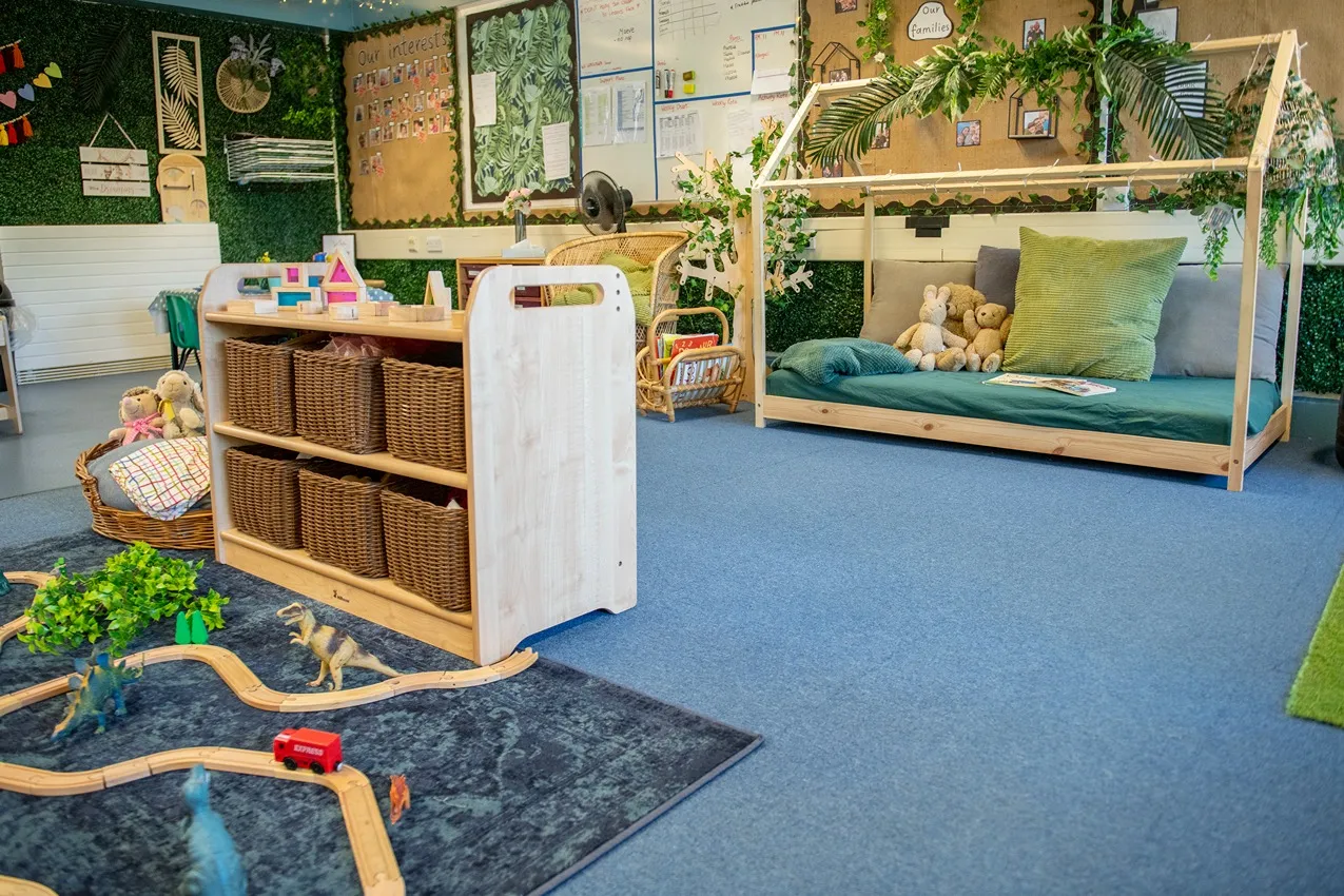 A bright nursery classroom featuring a wooden toy shelf, dinosaur train set, and cozy reading nook with a house frame bed.