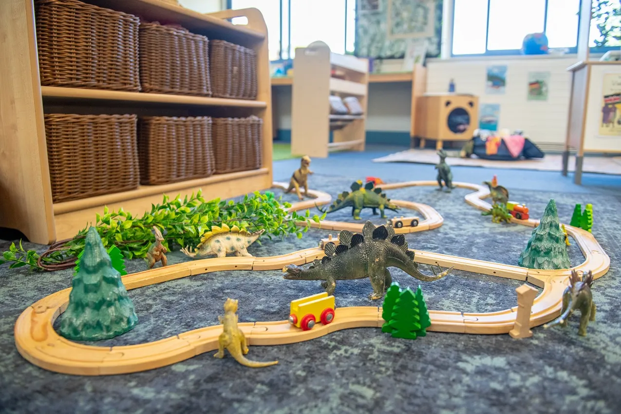 A wooden train track with toy dinosaurs and trees in a nursery playroom.