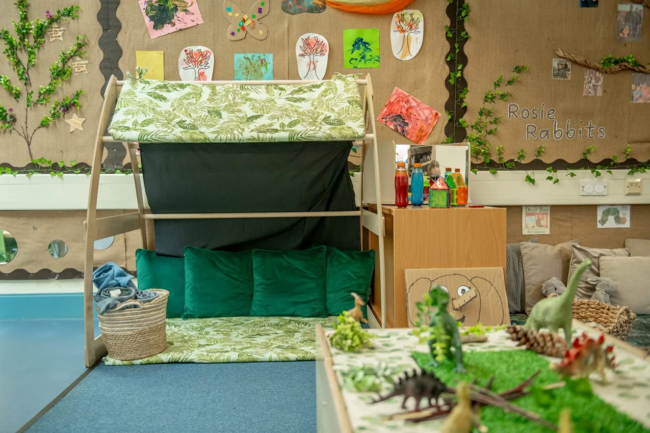 A cozy reading nook with green cushions and a leaf-patterned canopy in a bright nursery classroom.