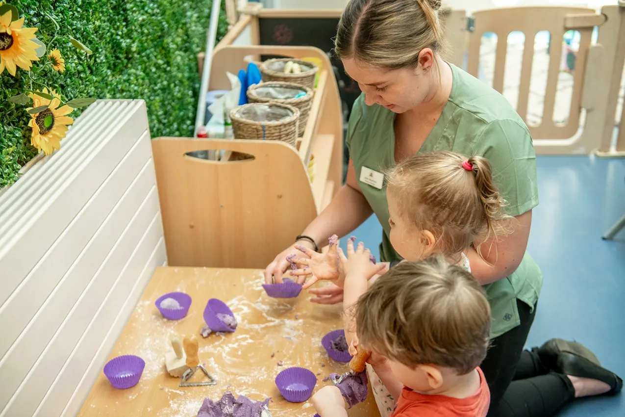 A nursery teacher helps toddlers with a purple playdough activity.