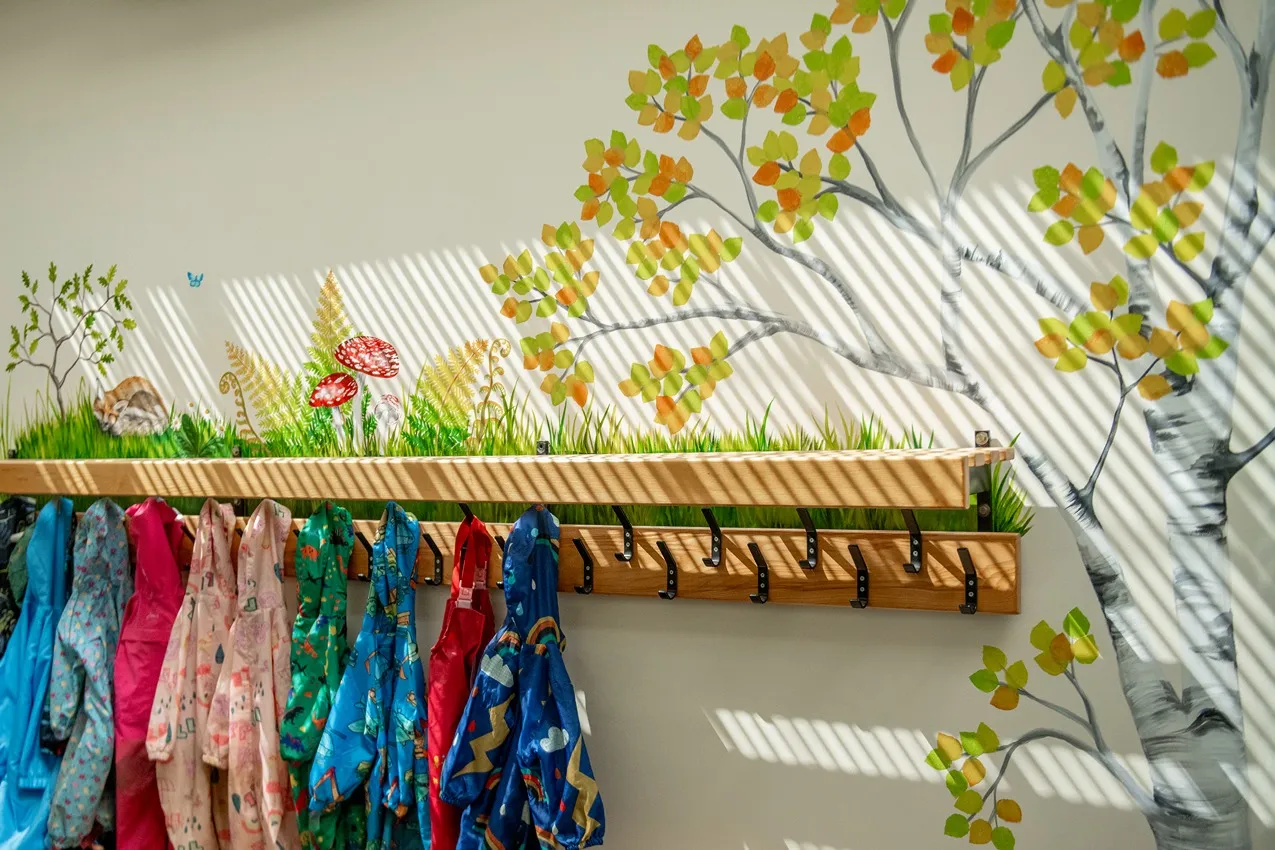 Colorful raincoats hang below a whimsical forest mural in a nursery school cloakroom.