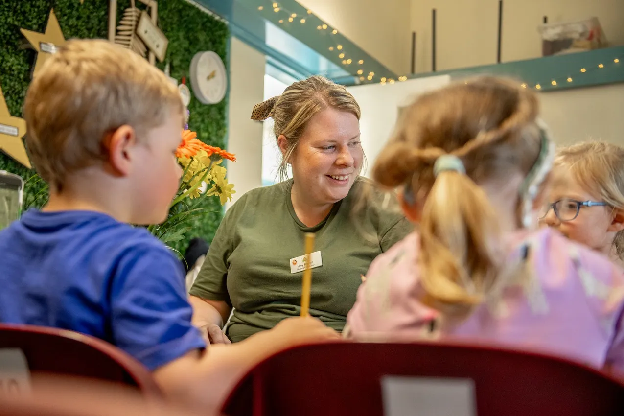 Children playing at a wooden play kitchen with a teacher in a bright nursery setting.