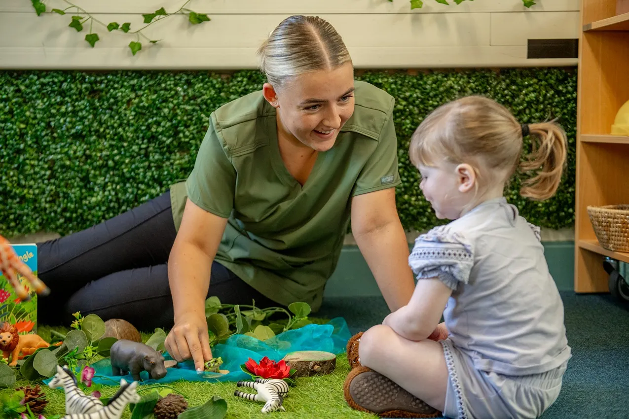 A nursery teacher and toddler play with toy animals in a jungle-themed learning environment.