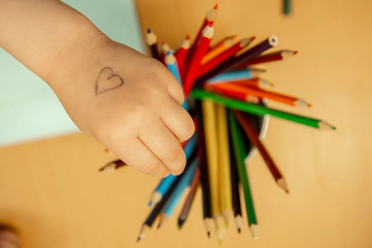 Child's hand with heart drawing reaching for colorful pencils. Nursery art supplies for creative learning.