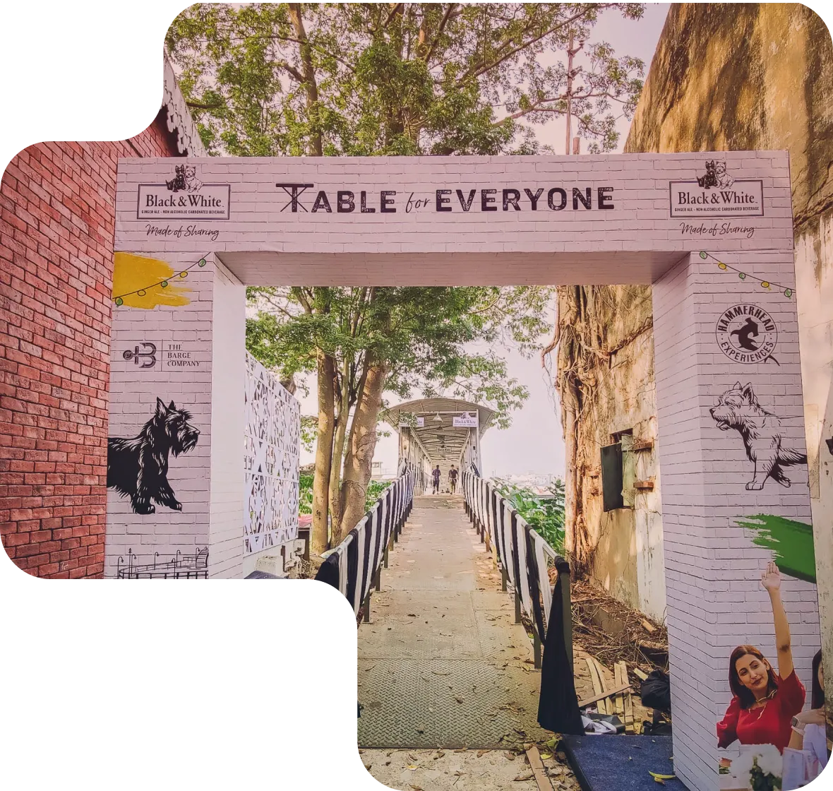 Entrance gate with 'Table for Everyone' text, decorated with dog illustrations and brand logos, leading to a walkway surrounded by trees.