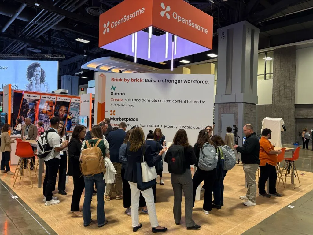 Group of attendees gathered at the OpenSesame booth at ATD 2025, with a large overhead sign and display reading “Brick by brick: Build a stronger workforce.” Promotional panels highlight offerings like “Simon” for custom content creation and “Marketplace” for curated courses.