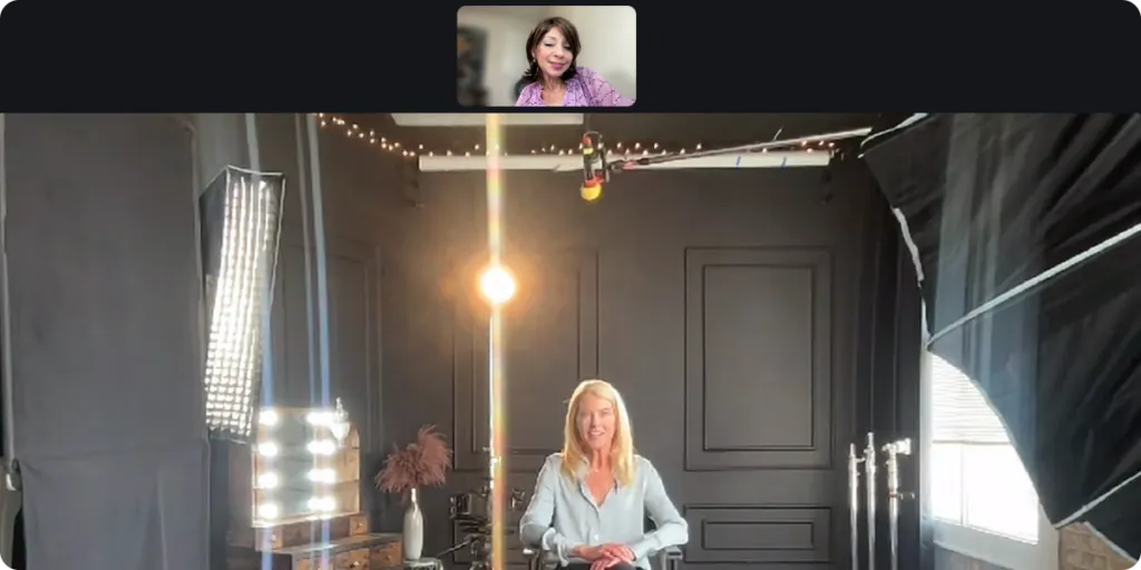 Karen Harding and Robin Hauser in a video call setup. Karen Harding in the smaller inset at the top is wearing a purple outfit and appears on a video call screen. The main scene shows Robin Hauser seated in a studio with black paneled walls, professional lighting equipment, and decorative string lights above.