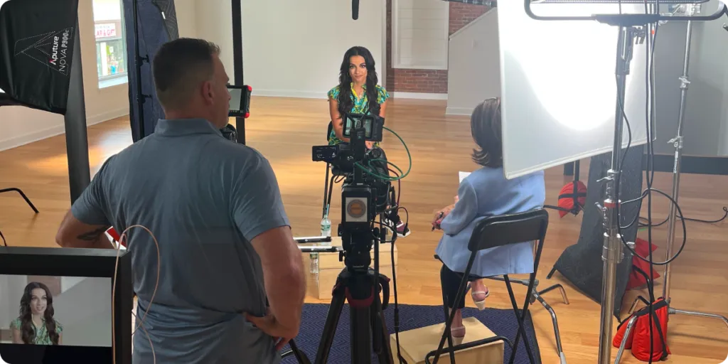Filming setup in a studio with a woman in a green top seated facing the camera, Karen Harding in a light blue blazer seated with her back to the camera, and a man operating filming equipment. Professional lighting, tripods, monitors, and cables are visible throughout the room with hardwood floors and white walls.