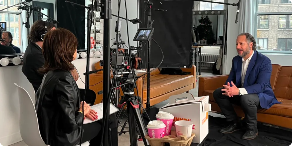 Interview setup in a studio with a man in a blue blazer seated on a brown couch, speaking to Karen Harding in a black leather jacket seated across from him. Camera equipment, lighting, and a monitor operator are visible. A Dunkin’ coffee box and multiple cups are on a table in the foreground. Large windows and vanity mirrors are in the background.