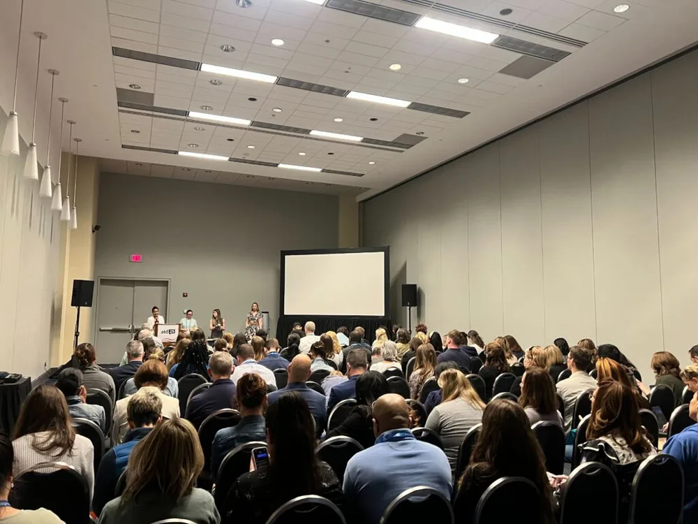 Conference room filled with seated attendees facing a panel of four speakers at the front near a podium and projection screen. The podium displays the ATD25 logo. This is a customer panel session.