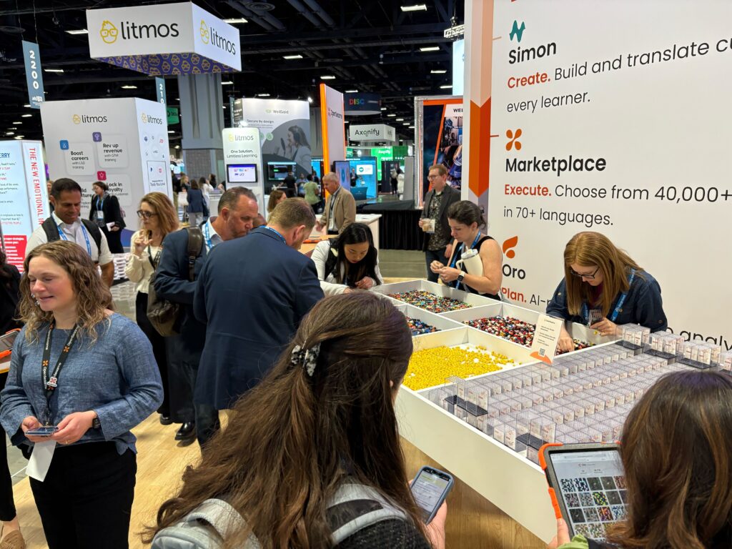 Attendees at the OpenSesame booth browsing bins of colorful LEGO bricks arranged in trays on a table, beneath a display highlighting products like Simon, Marketplace, and Oro. Nearby, other vendor booths are visible in the background.