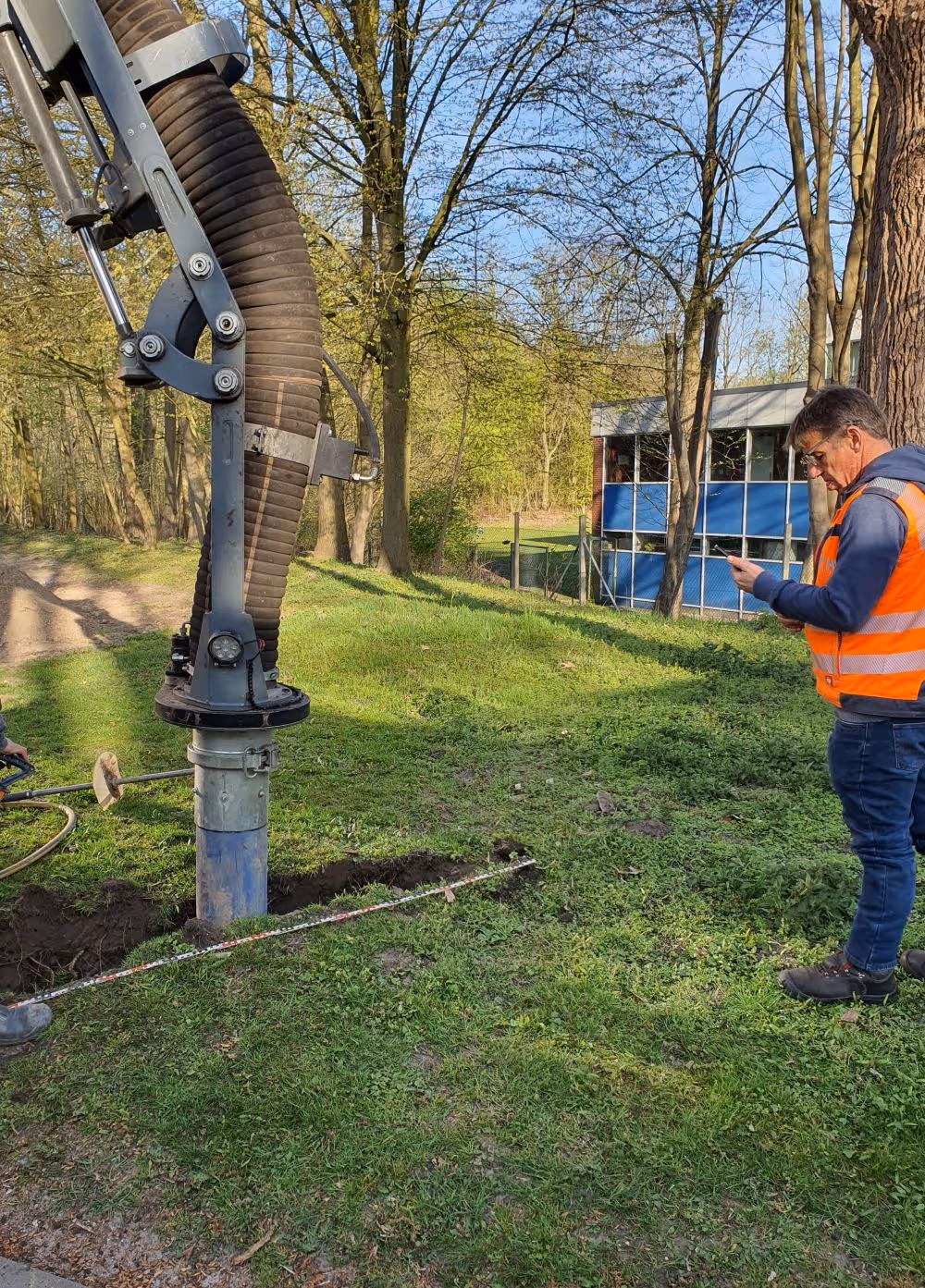 Mann in orangefarbener Sicherheitsweste steht auf Grasfläche neben einem großen Saugbaggerrohr nahe eines Gebäudes in einem bewaldeten Gebiet.