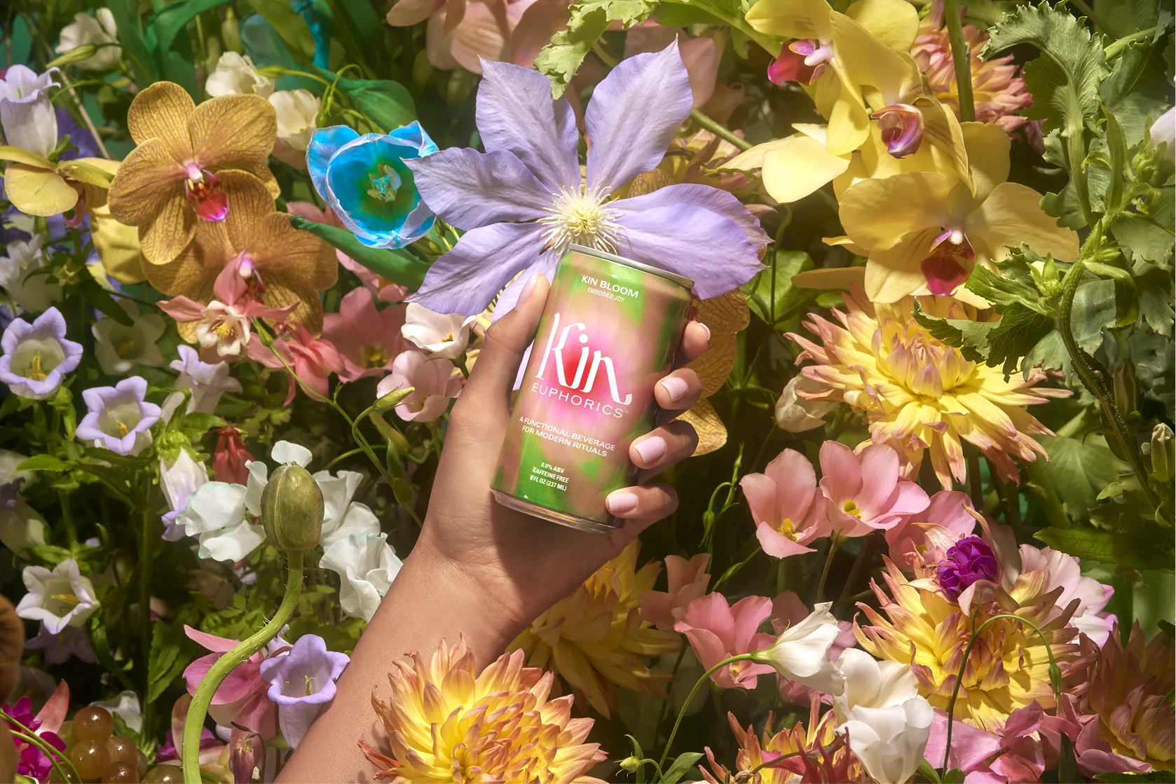 A hand holds a can of Kin Euphorics' "Kin Bloom" beverage, emerging from a dense background of colorful flowers.
