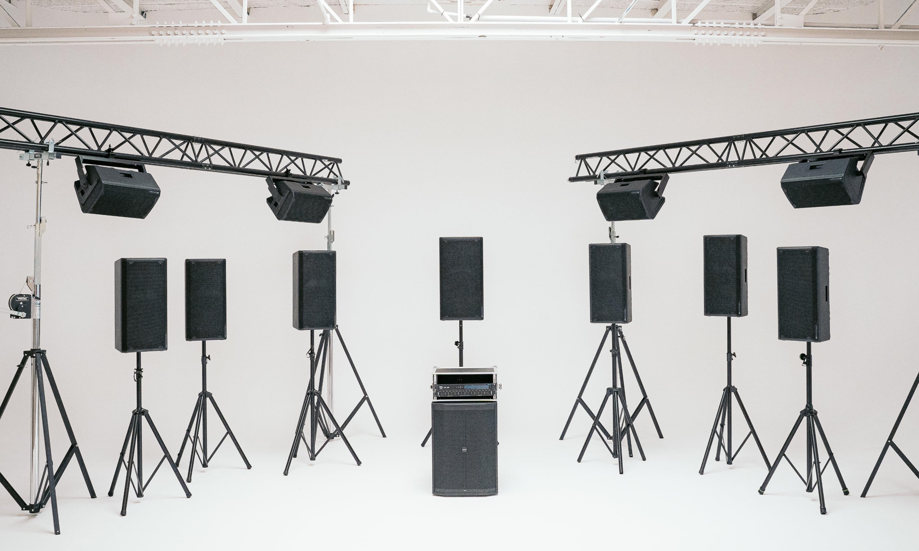 Studio setup with eight black speakers on tripods and two overhead lights on metal trusses against a white background.