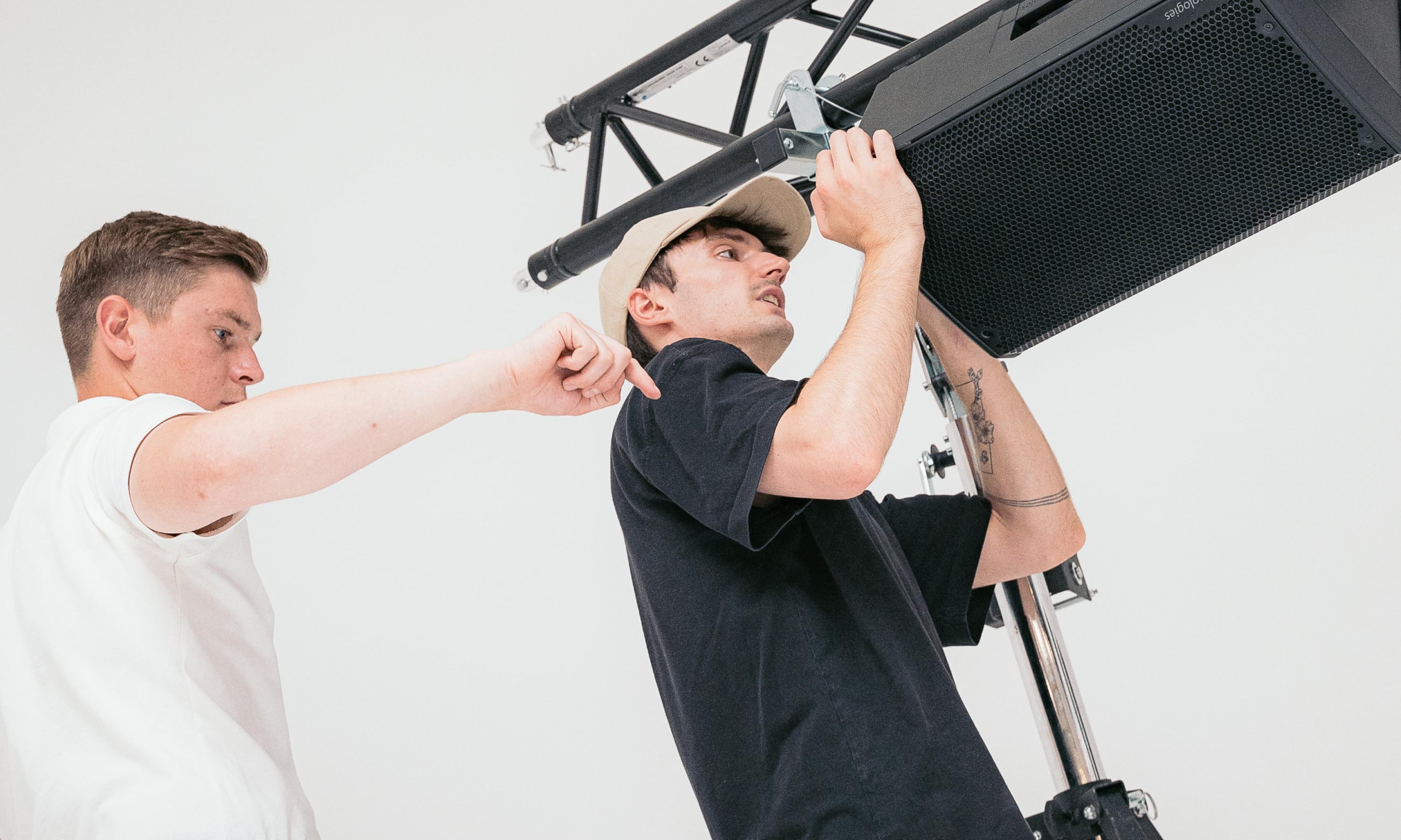 Two men adjusting a mounted black speaker on a metal stand against a white background.
