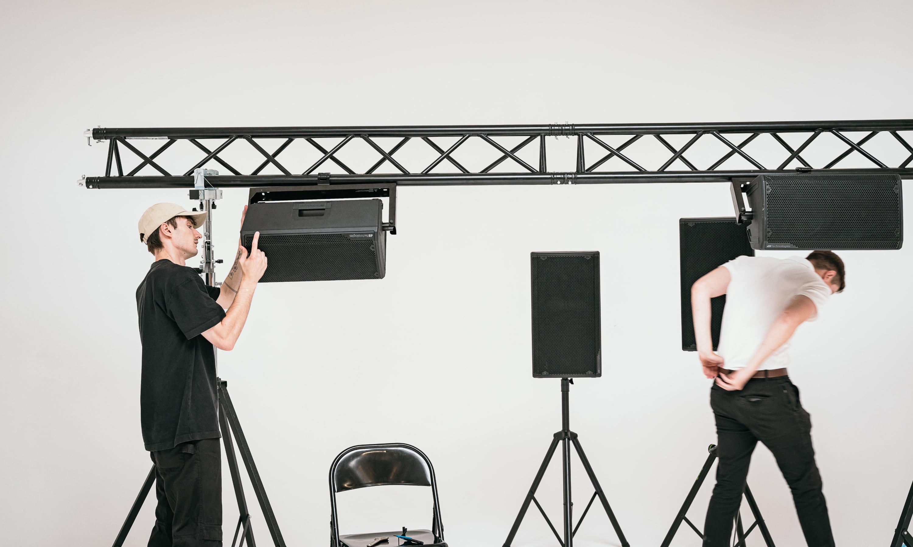 Two men setting up black speakers mounted on stands and a horizontal metal truss against a white background.