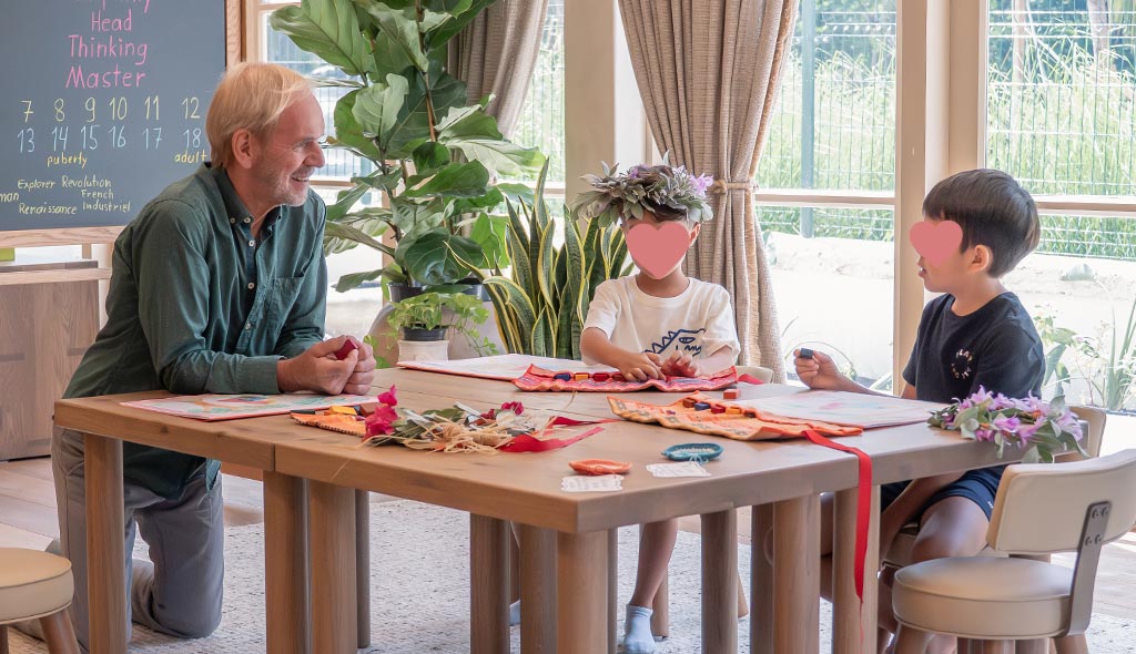 Older man kneeling by a table with two children seated, engaging in a learning activity with educational materials and plants in a light-filled room.