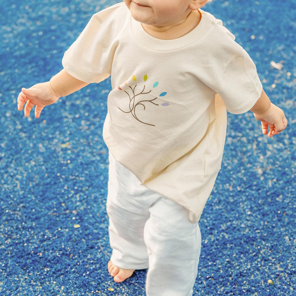 Toddler with bare feet standing on a blue speckled surface, wearing a cream shirt with a colorful tree design and white pants.