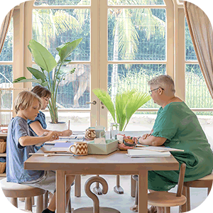 Two children and an older woman sitting at a wooden table studying or doing homework in a bright room with large windows and plants.