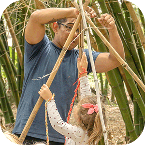 Man helping a child build a structure with bamboo sticks outdoors surrounded by bamboo plants.