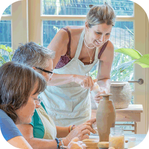 Three people engaged in pottery making with a tall clay vase on a wooden table in a sunlit room.