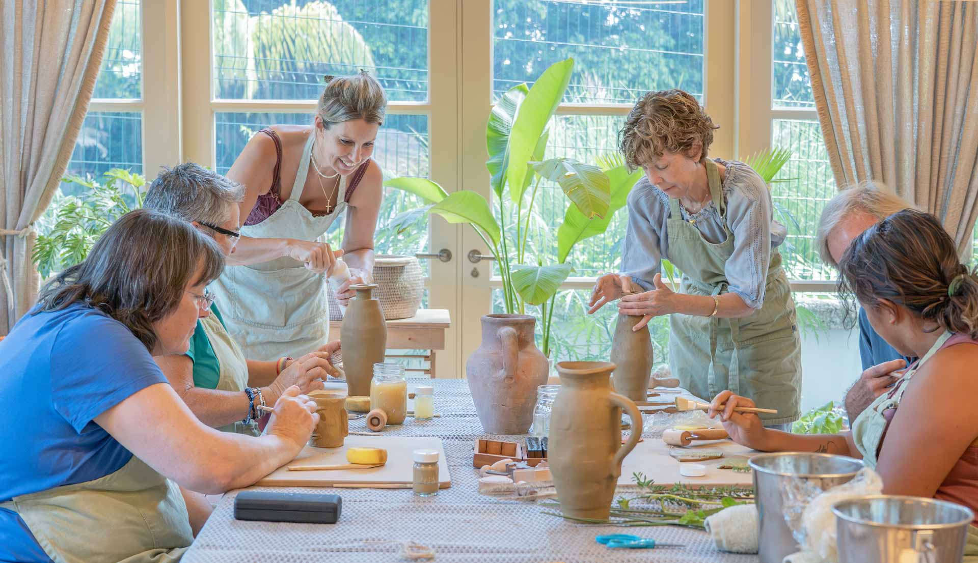 Six adults sitting around a table engaged in pottery crafting and painting in a bright room with large windows and green plants outside.
