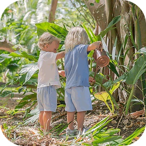 Two young children watering plants in a garden surrounded by greenery.