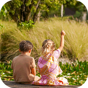 Two children sitting on a wooden surface outdoors, one holding a tall grass stalk raised in the air, surrounded by greenery.