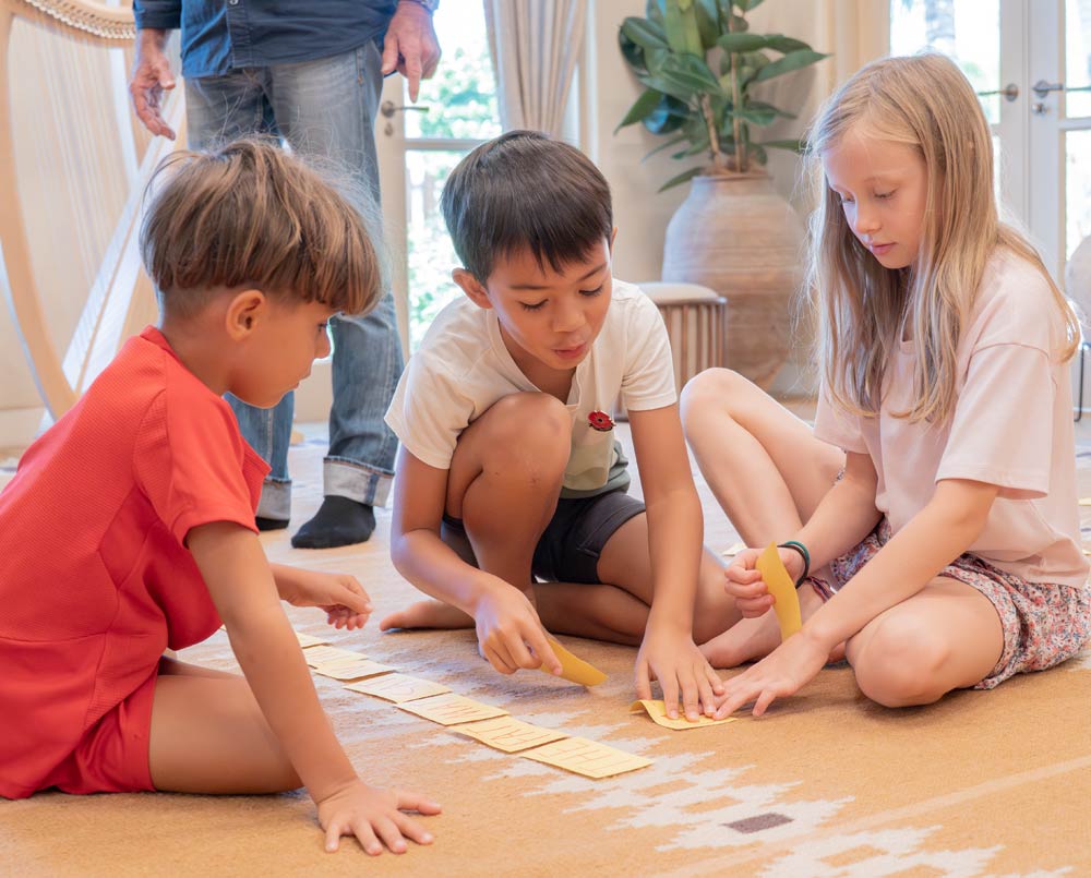 Three children sitting on a carpet, engaging with yellow paper strips arranged in a line, with an adult standing in the background.