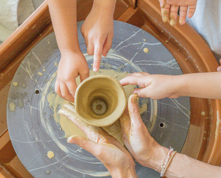 Hands shaping a clay pot on a pottery wheel, with adult and child hands working together.