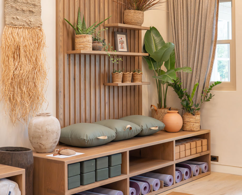 Well-organized yoga studio corner with cushions, yoga mats, blocks, and potted plants on wooden shelves and bench.
