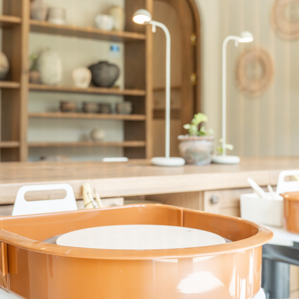 Close-up of a brown pottery wheel in a bright room with wooden shelves holding pottery in the background.