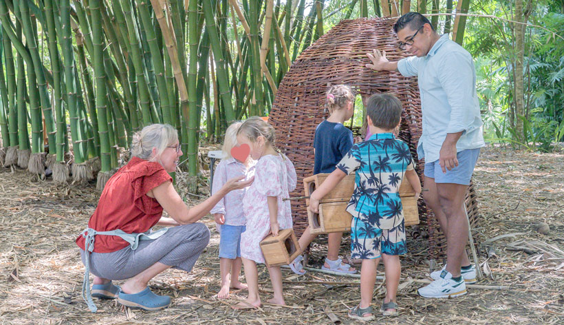 An adult woman crouching and interacting with three children holding wooden blocks, while another adult man stands nearby, outside near bamboo and a woven structure.