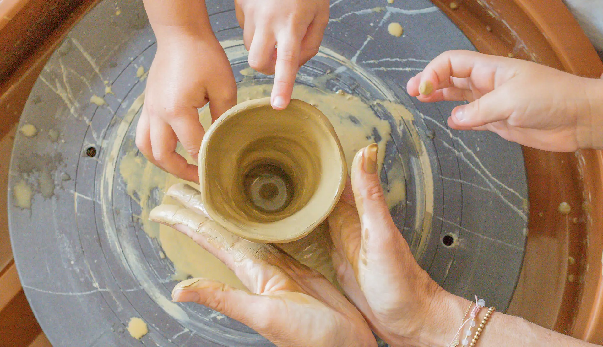 Hands shaping a clay pot on a pottery wheel with assistance and clay-covered fingers.