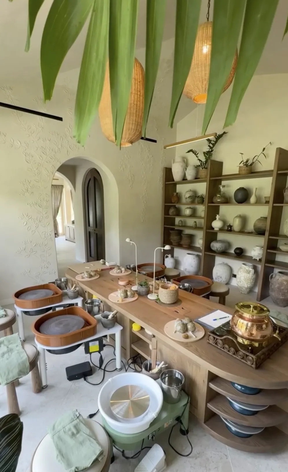A pottery studio with four pottery wheels arranged around a wooden table. The table holds several pieces of clay work and pottery tools. Shelves in the background display various ceramic pots and vases. Large green leaves partially frame the upper part of the image, and woven pendant lights hang from the ceiling.