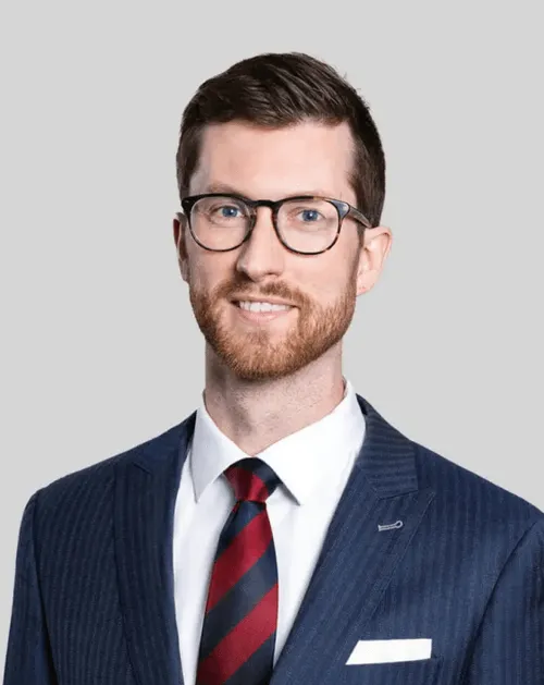 Smiling man with glasses, brown hair, and beard wearing a navy pinstripe suit, white shirt, and red and black striped tie.
