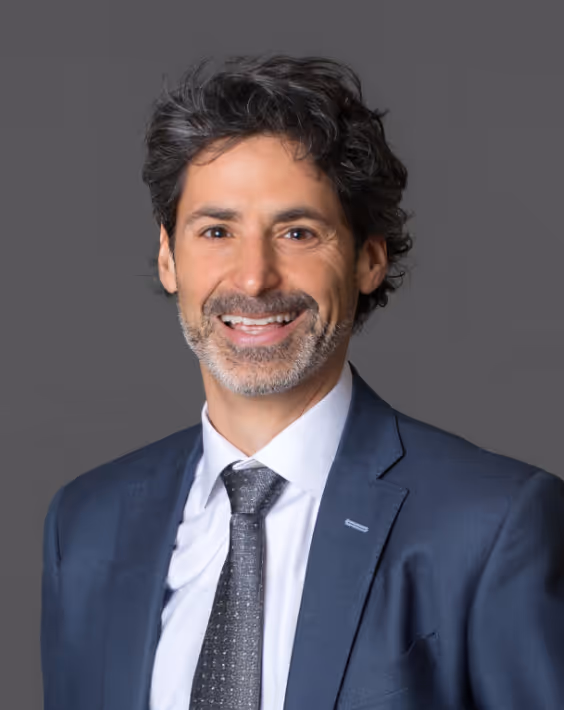 Smiling man with dark curly hair and beard wearing a dark blue suit, white shirt, and gray tie against a gray background.