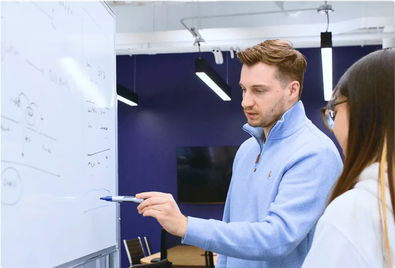 Man in light blue sweater explaining concepts on a whiteboard to a woman in an office setting.