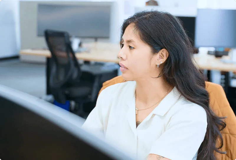 Young woman with long dark hair wearing a white shirt, sitting at a desk in an office and looking to the side.