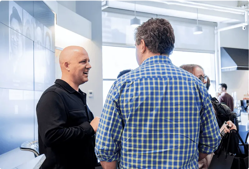 Two men engaged in conversation in a modern office setting, one wearing a black shirt and the other a blue checkered shirt.