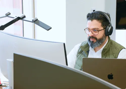 Man with glasses and beard wearing headphones and a green vest working at a desk with multiple computer screens and a laptop.