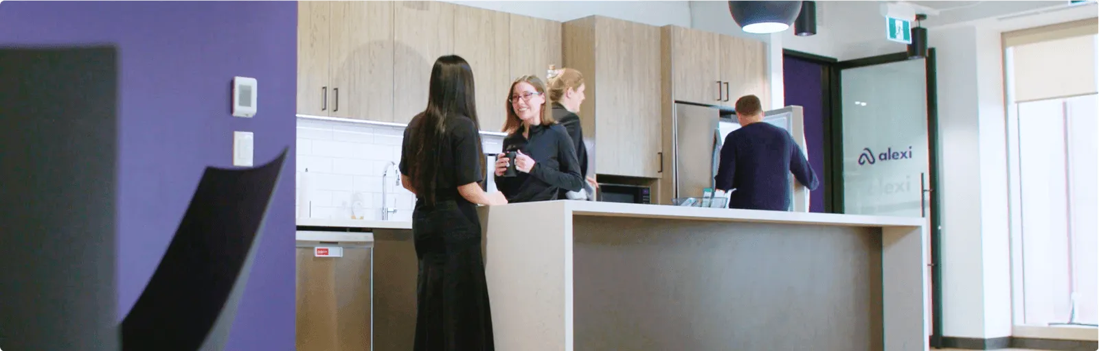 Office kitchen area with three people conversing near the counter and one person opening a fridge in the background.