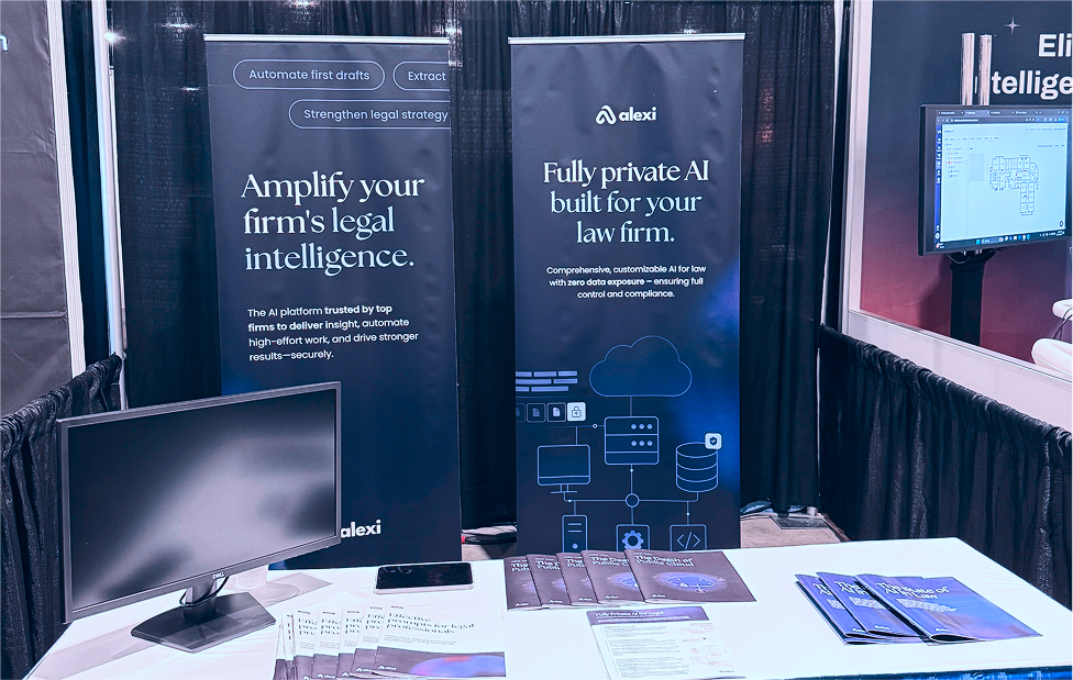 Trade show booth with two vertical banners promoting Alexi's AI legal tech, a blank Dell monitor on a table with brochures and flyers.