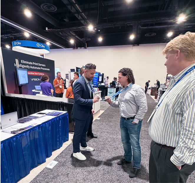 Three men conversing in a conference exhibit hall with booths displaying technology solutions.
